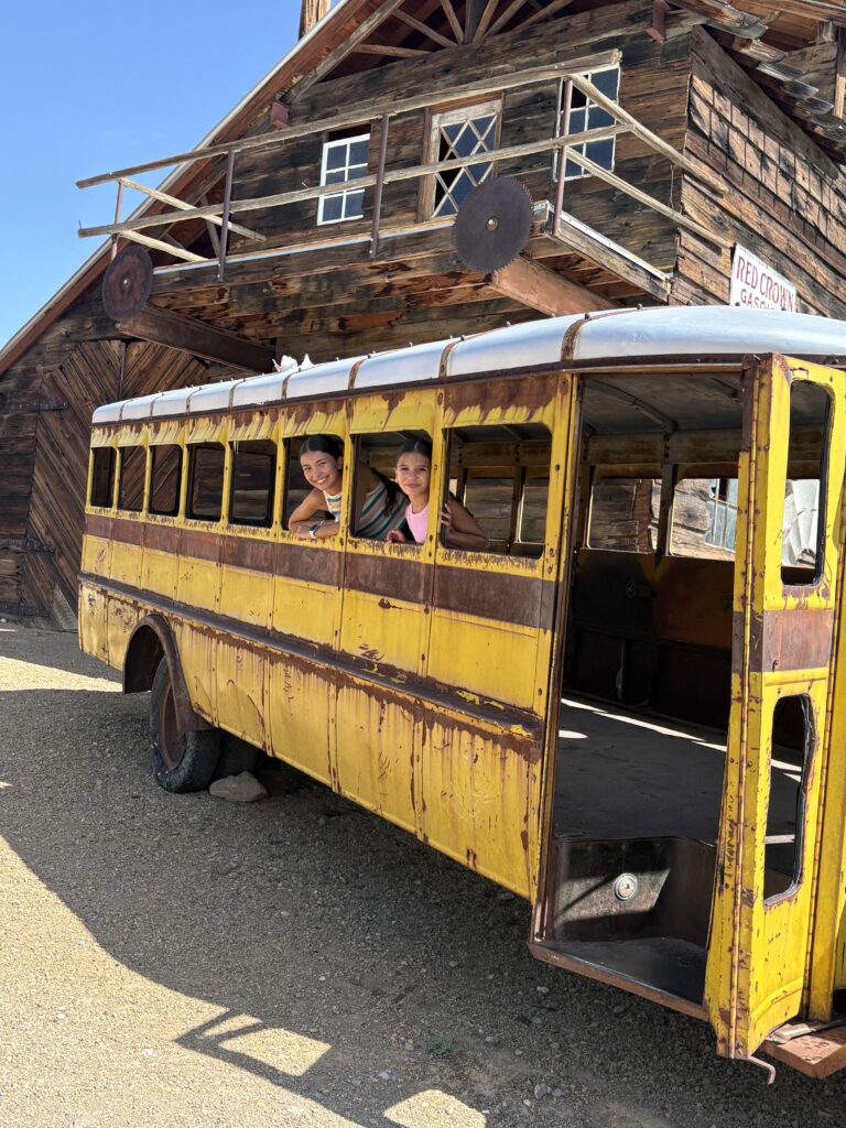 Two smiling children look out from a yellow bus in front of a rustic Nevada ghost town near the Colorado River under blue skies.