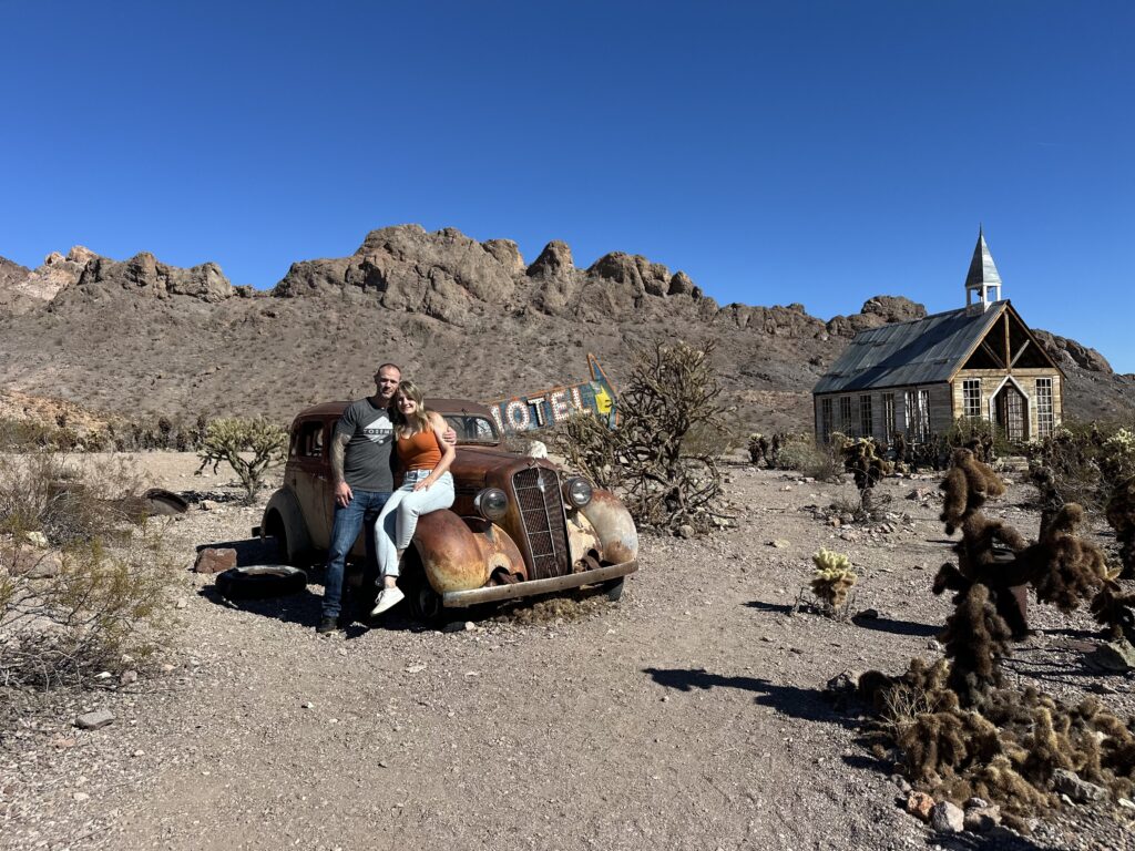 Couple poses by rusty vintage car at Nelson Ghost Town near Las Vegas, Nevada, with rocky hills and historic sights in the background.