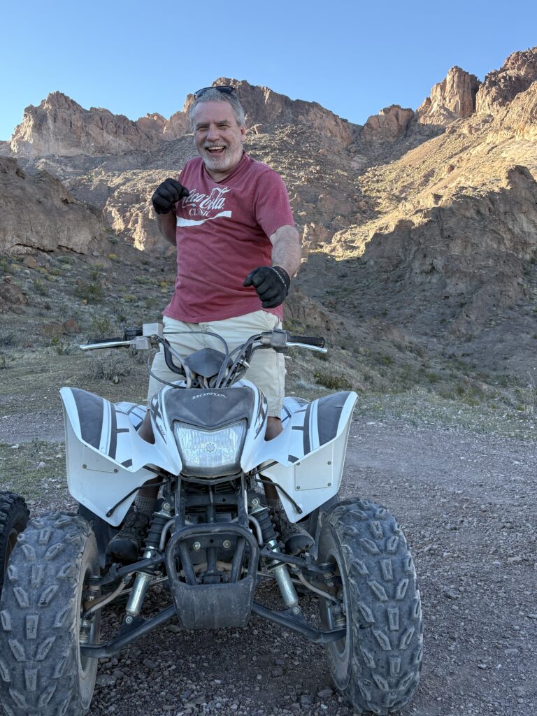 Smiling man in red tee by white ATV on rocky Nevada trail—Las Vegas ATV tour near desert mountains, clear sky in background.