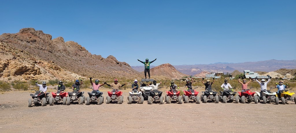 Group on ATVs poses with arms raised on a Nevada desert ATV tour near Las Vegas, rocky hills and blue sky in the background.