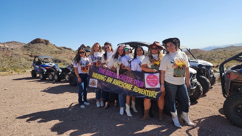Eight women on a Nevada ATV tour near Las Vegas smile by desert ATVs, holding “The Bach Lorettes Trip” adventure banner.